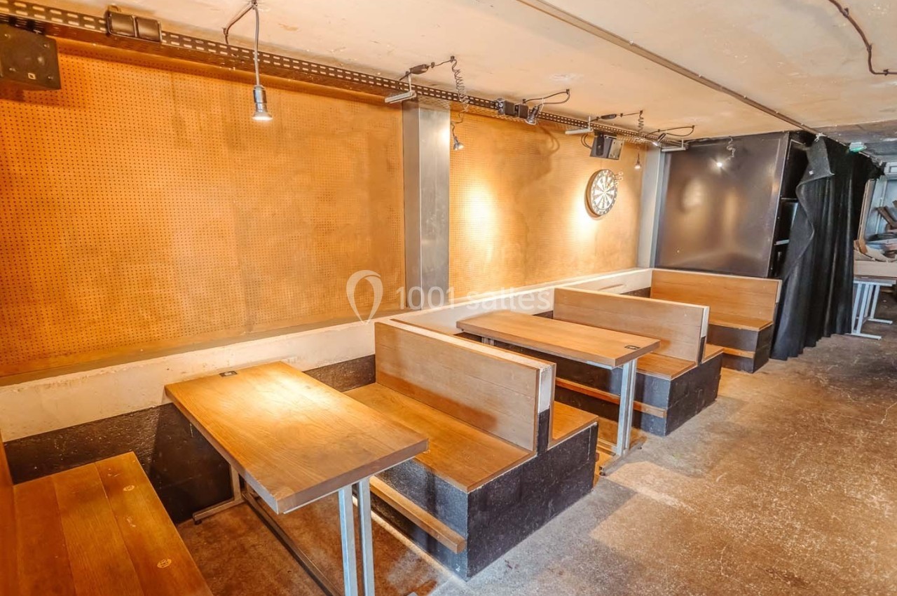 Banquettes en bois alignées le long d'un mur beige avec un jeu de fléchettes accroché, dans un espace lumineux.