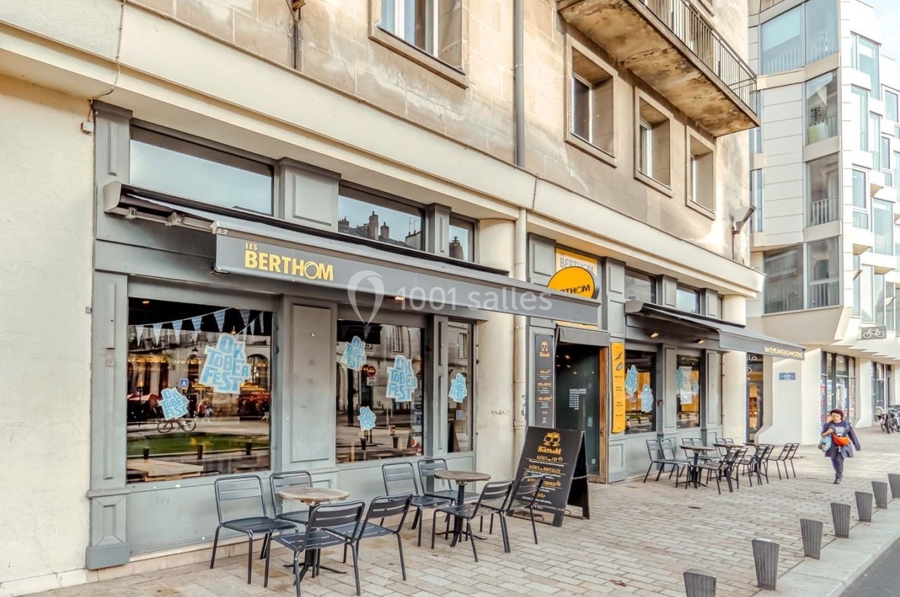 Façade d'un bar avec terrasse extérieure, tables et chaises, situé dans une rue bordée de bâtiments modernes et anciens.