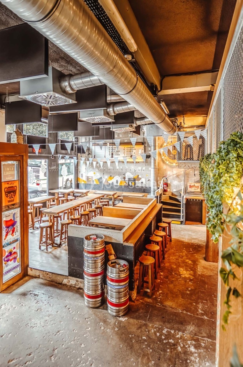 Intérieur d'un bar avec tables en bois, fûts de bière, guirlandes décoratives et lumière naturelle.