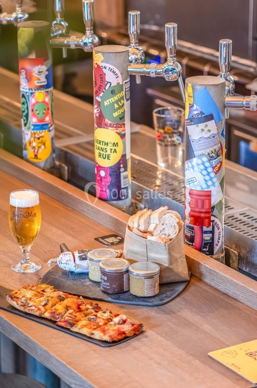 Planche apéritive avec tartines et condiments, accompagnée d'une bière, posée sur un comptoir de bar avec tireuses colorées.