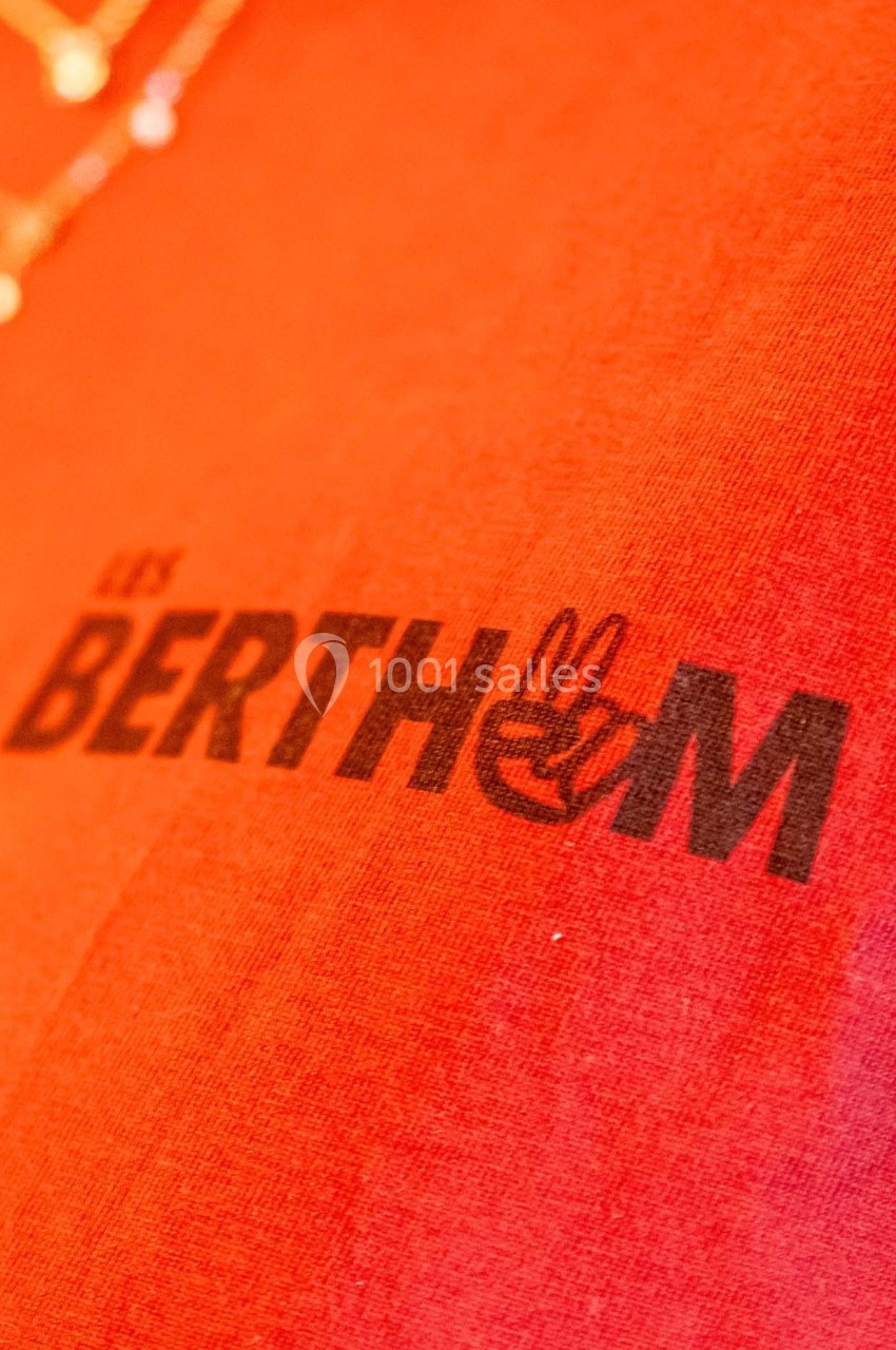 Logo ’Les Berthom’ noir sur un tissu rouge avec un motif de main formant un signe de paix dans la lettre ’M’.