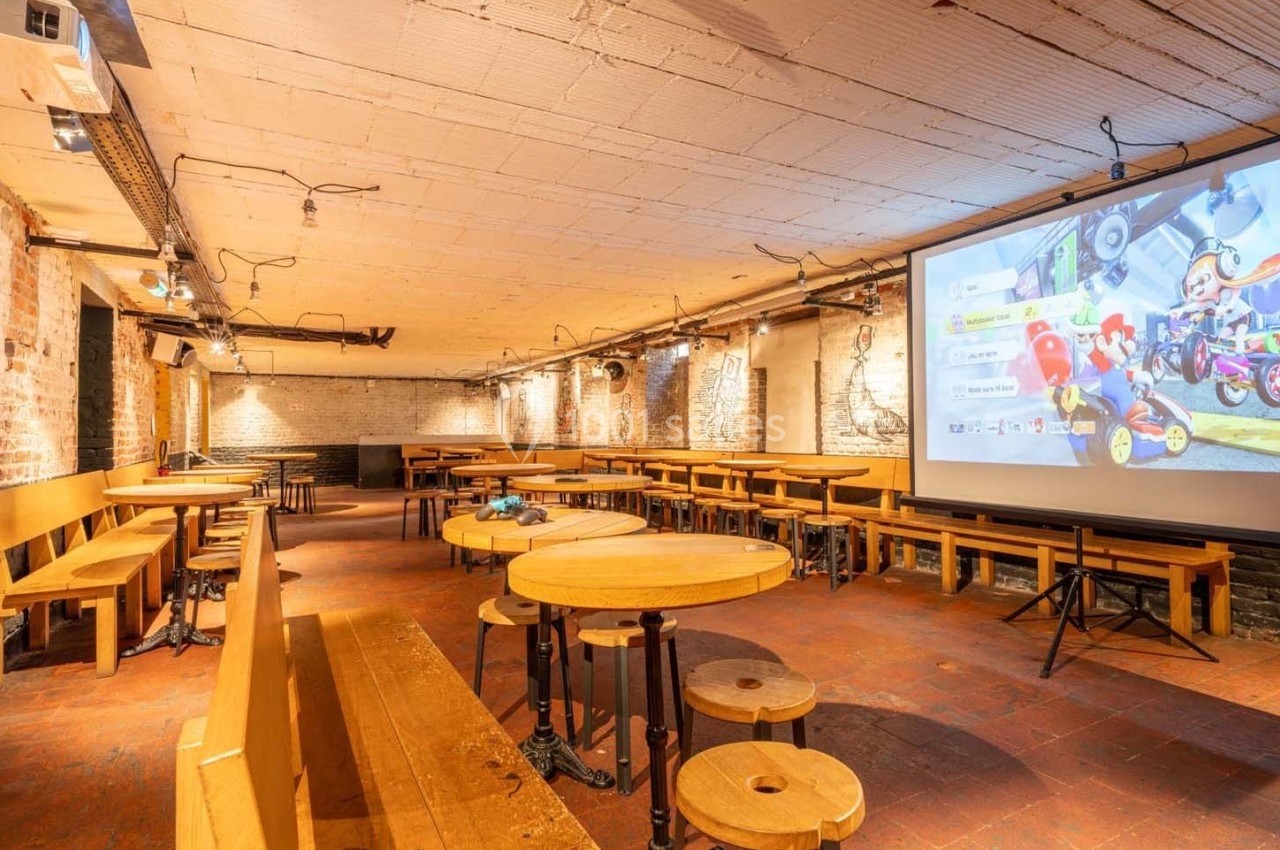 Salle voûtée avec murs en briques, tables en bois et écran géant affichant un jeu vidéo.