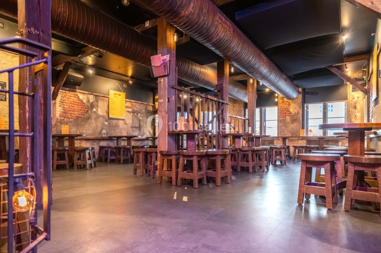 Salle de restaurant vide avec tables et tabourets en bois, murs en briques et tuyaux apparents au plafond.