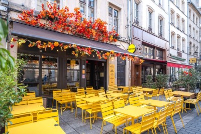 Terrasse d'un café avec des chaises jaunes, des clients attablés et une façade décorée de feuillage automnal.