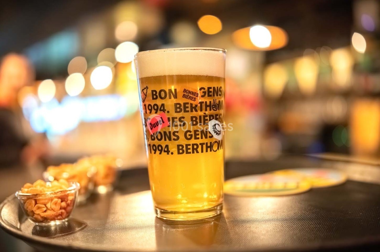 Verre de bière blonde sur un plateau avec des cacahuètes en arrière-plan flou dans un bar éclairé.