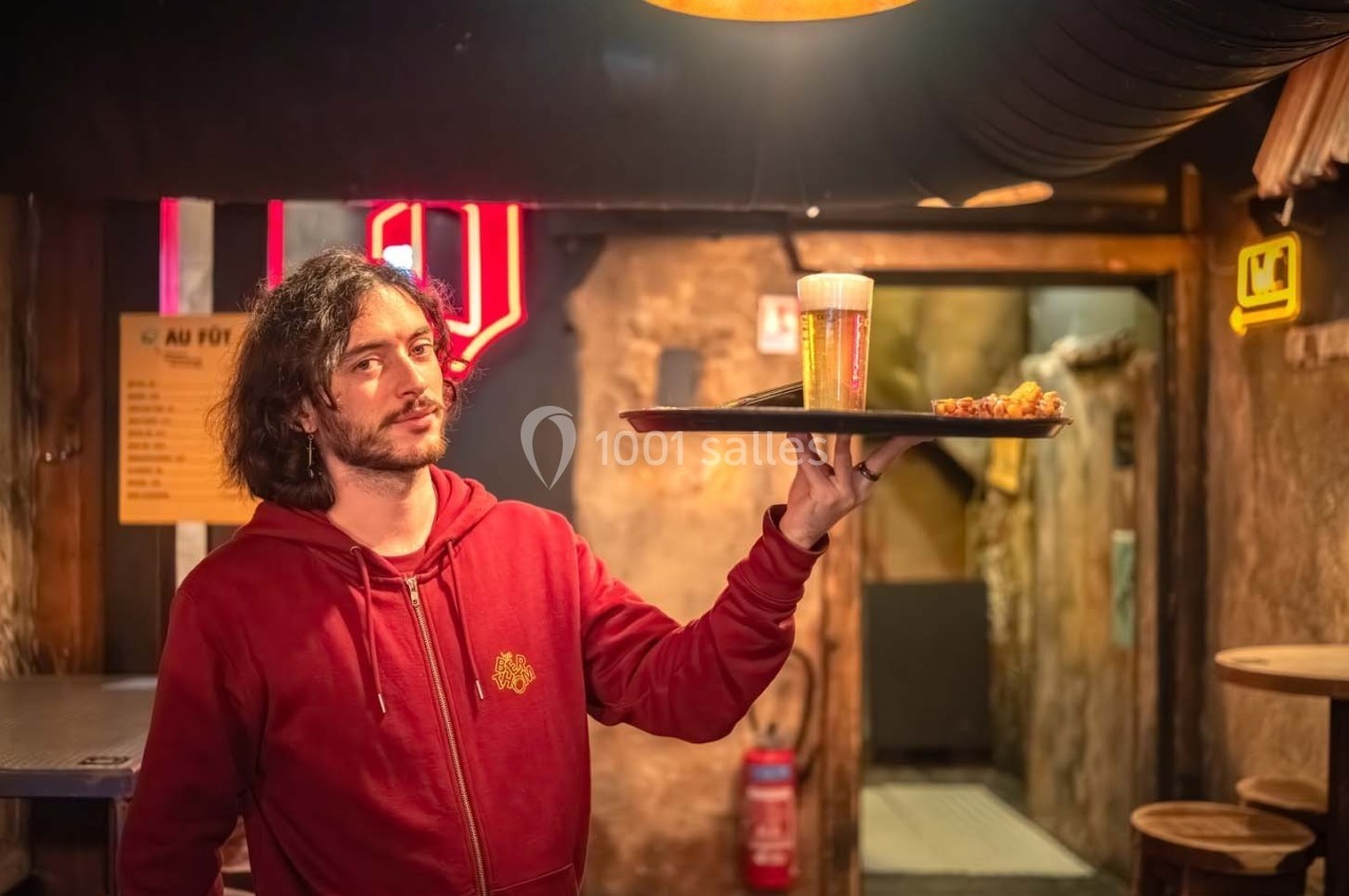 Un serveur tenant un plateau avec une bière et un plat dans un bar à l'ambiance rustique et éclairage chaleureux.