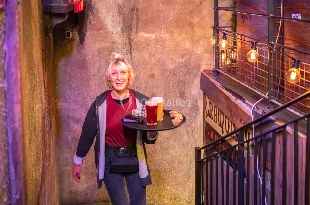 Serveuse souriante portant un plateau avec des boissons et des snacks dans un escalier éclairé par des guirlandes lumineuses.