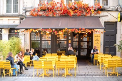 Terrasse d'un café avec des chaises jaunes, des clients attablés et une façade décorée de feuillage automnal.