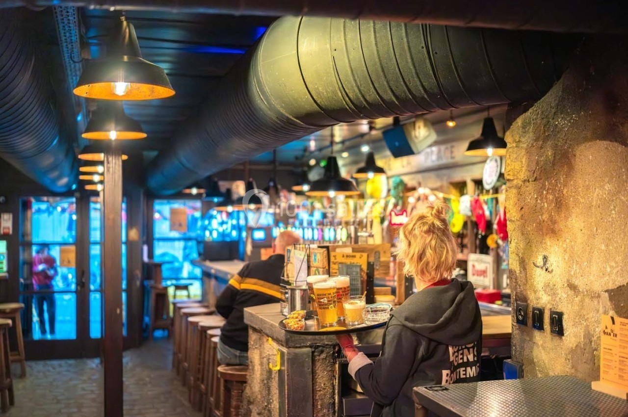 Intérieur d'un bar chaleureux avec un comptoir en bois, des tabourets alignés et une serveuse portant des boissons.