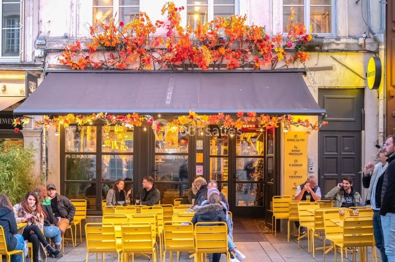 Terrasse d'un café avec des chaises jaunes, des clients attablés et une façade décorée de feuillage automnal.