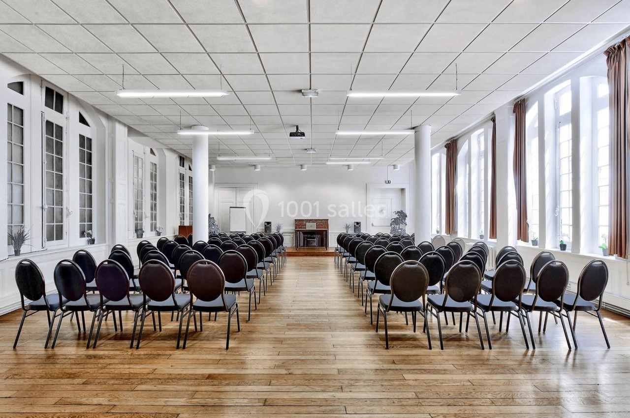 Salle lumineuse avec parquet, rangées de chaises noires alignées face à un bureau et un mur de fenêtres.