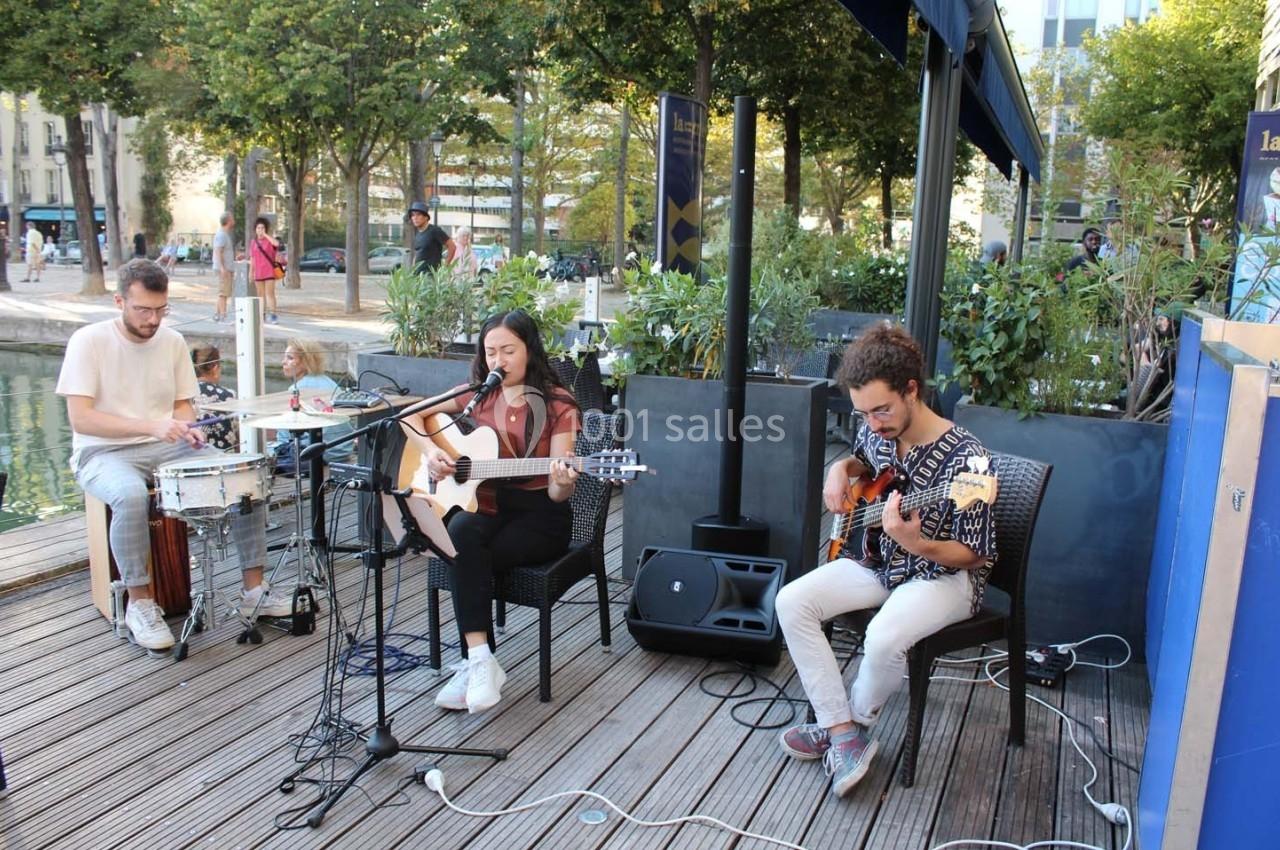 Un groupe de musiciens joue en plein air sur une terrasse en bois, avec une chanteuse à la guitare au centre.
