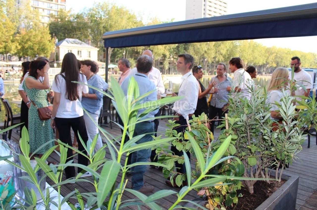 Groupe de personnes discutant sur une terrasse en bois avec des plantes au premier plan et des arbres en arrière-plan.