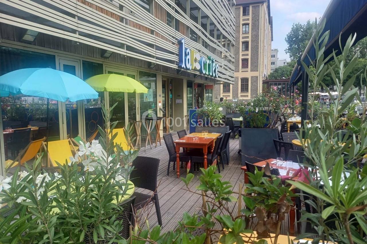 Terrasse d'un café avec tables, chaises colorées, parasols et plantes, située devant un immeuble moderne.