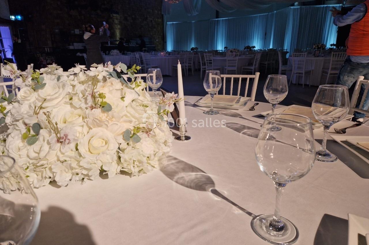 Table dressée avec une nappe blanche, centre de table floral, verres à vin et chandelles dans une salle décorée.