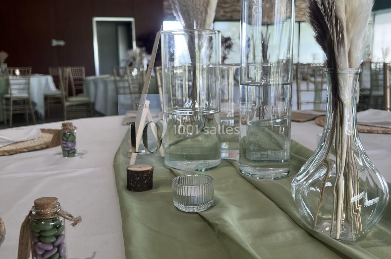 Centre de table avec vases en verre, herbes séchées et décorations sur une nappe verte dans une salle de réception.