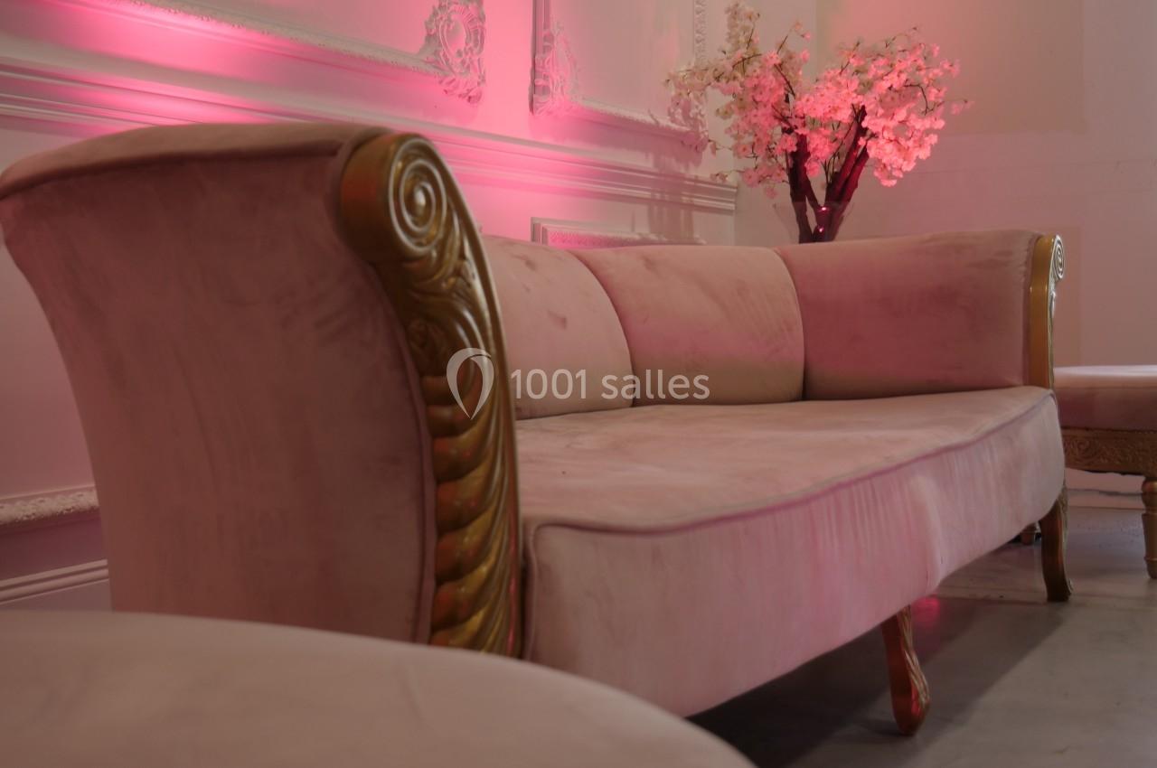 Canapé en velours rose avec accoudoirs dorés, placé dans une pièce ornée de moulures et éclairée par une lumière rose.