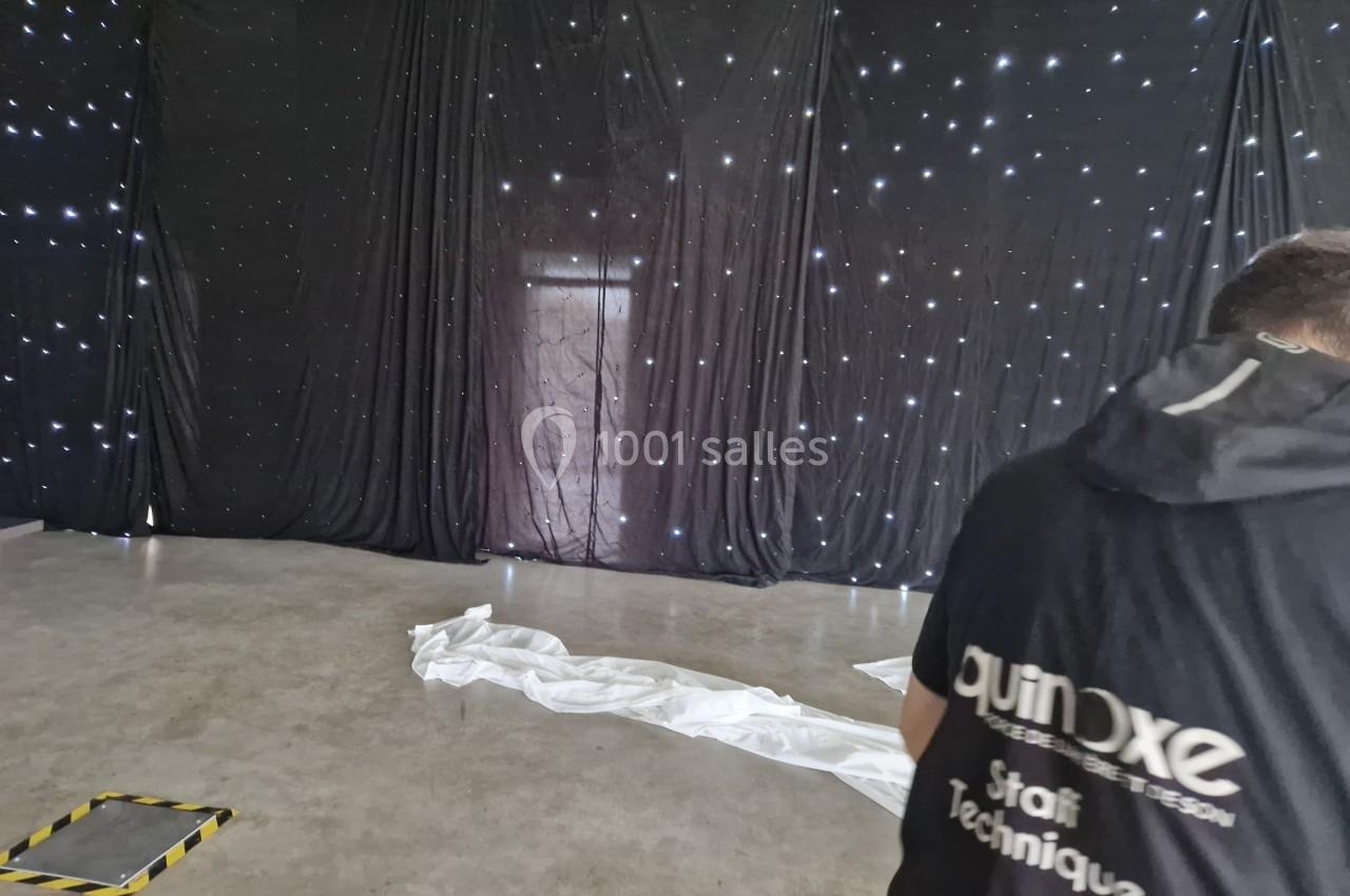 Rideau noir étoilé en fond, sol en béton avec tissu blanc au sol, personne de dos portant un vêtement marqué ’staff…