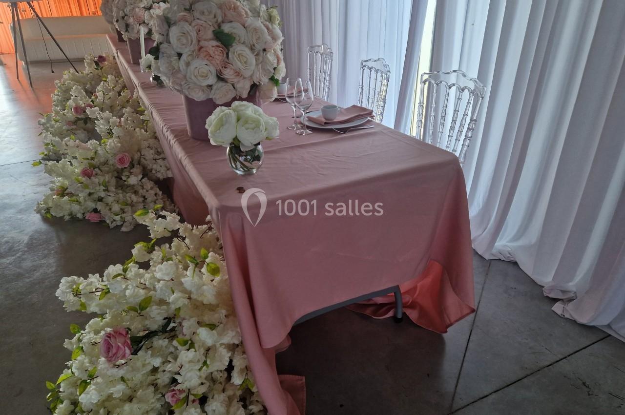 Table décorée avec une nappe rose, des bouquets de fleurs blanches et roses, et des chaises transparentes.