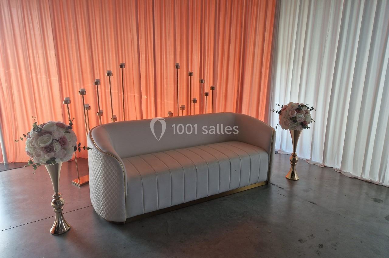 Canapé blanc devant un rideau orange et blanc, encadré par deux bouquets de fleurs et des bougeoirs dorés.