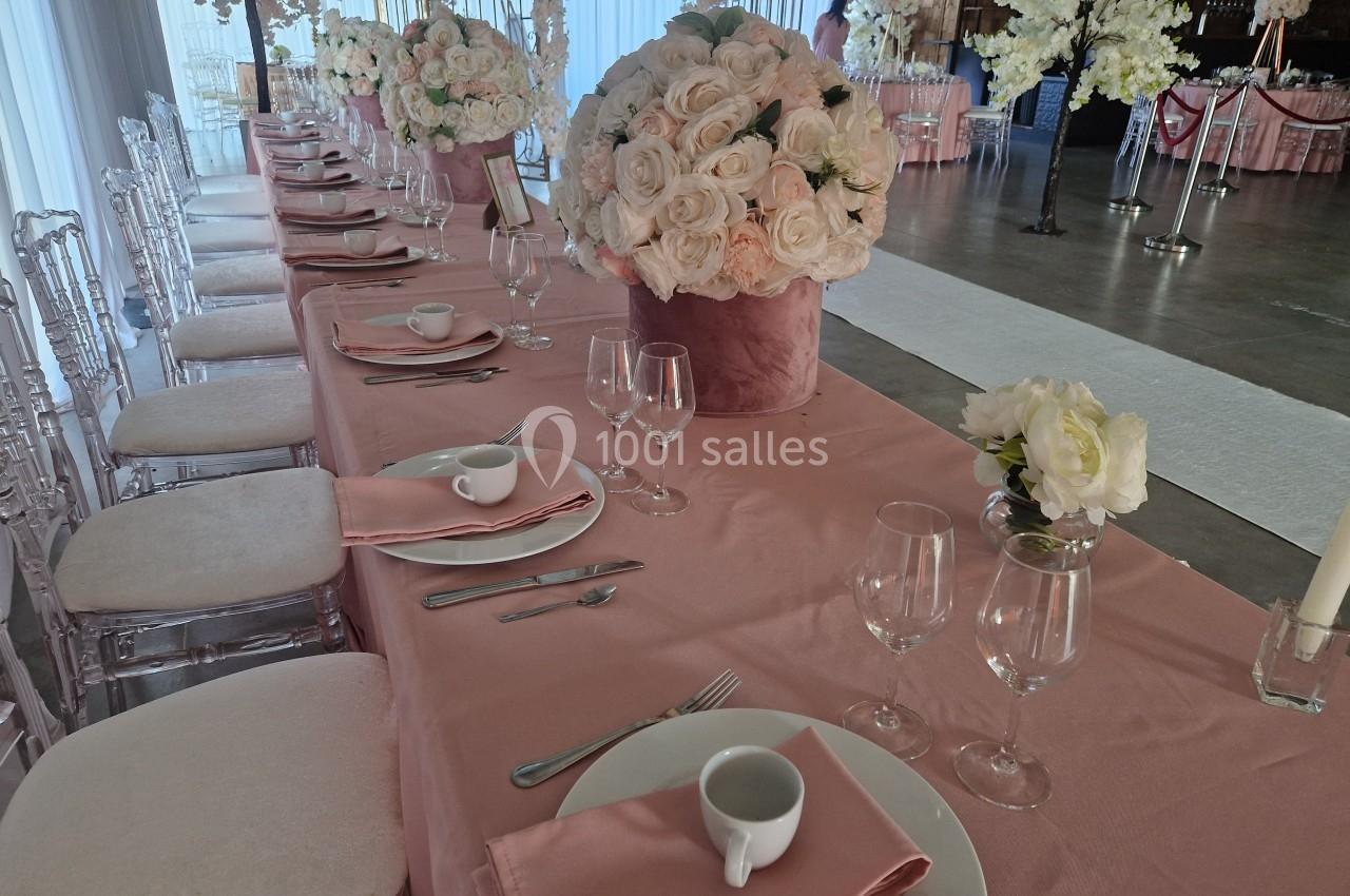 Table de réception élégante avec nappes roses, vaisselle blanche, verres et bouquets de roses blanches en centre de table.
