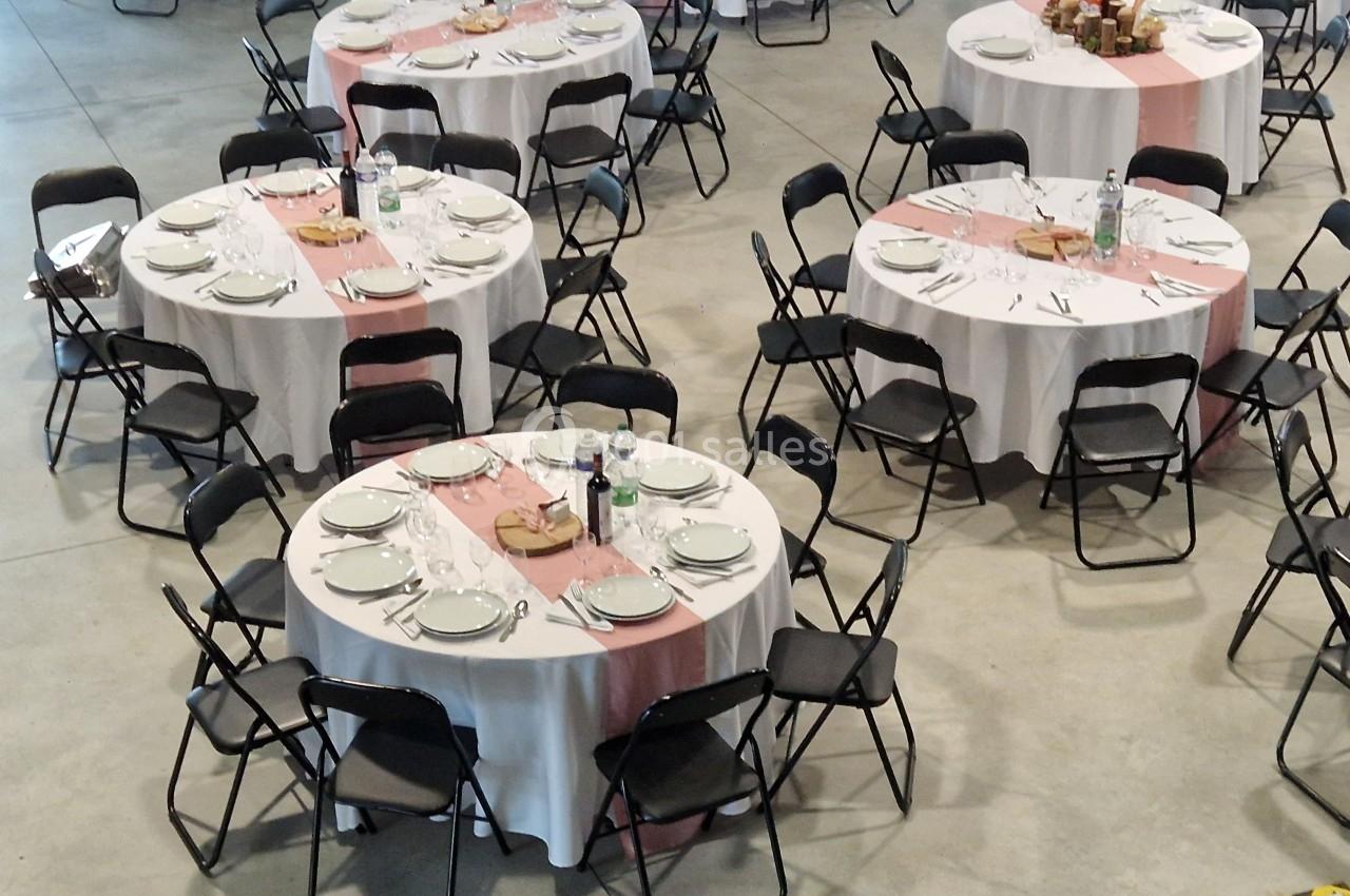 Tables rondes dressées avec nappes blanches, chemins de table roses, vaisselle et chaises noires dans une salle.