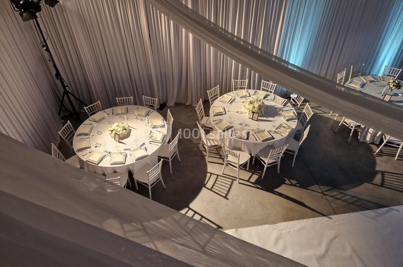 Tables rondes dressées avec nappes blanches, chaises assorties et centres de table fleuris dans une salle décorée de rideaux…