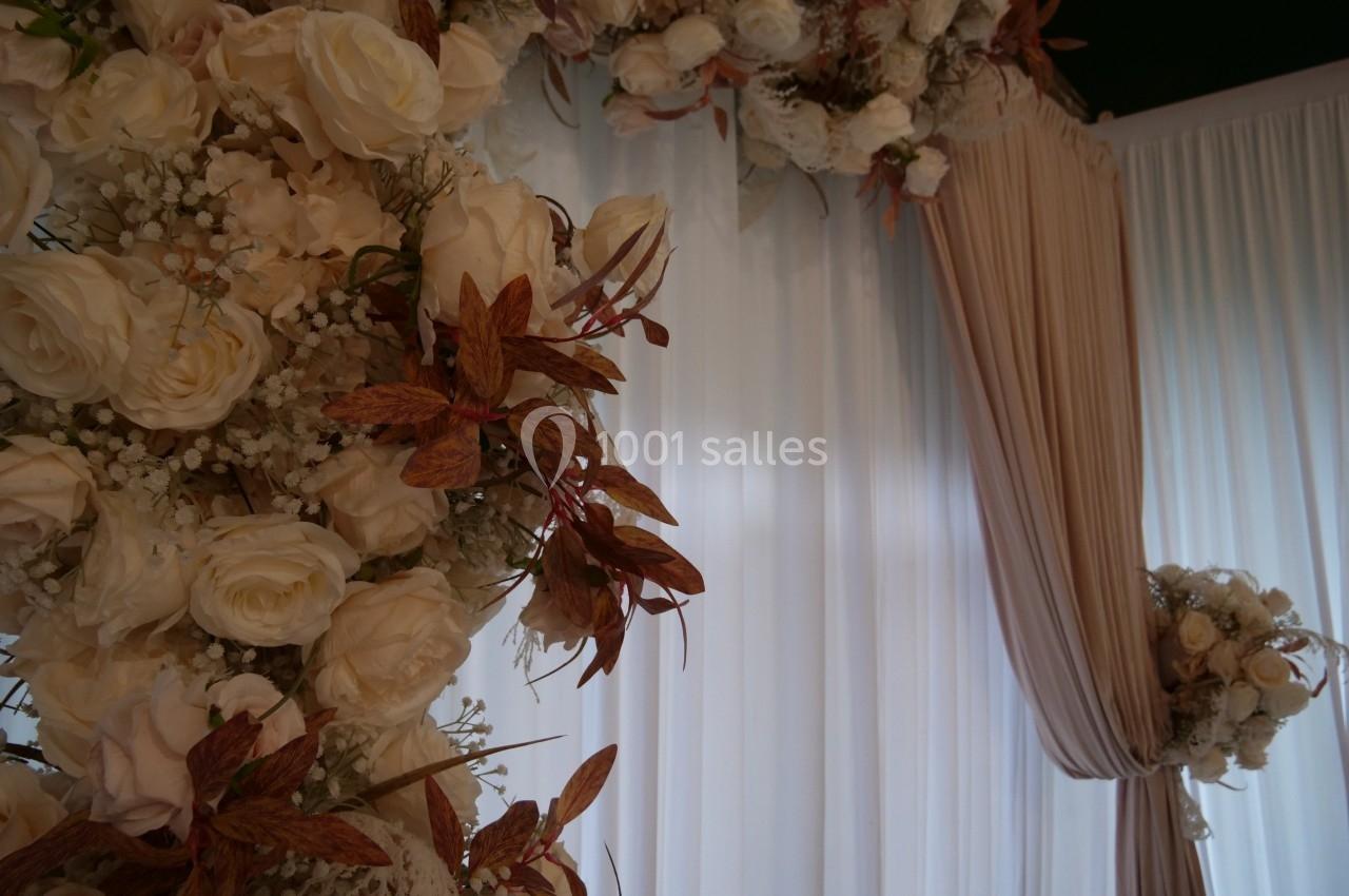 Arrangement floral avec des roses blanches et feuillage brun devant des rideaux blancs et beiges drapés.