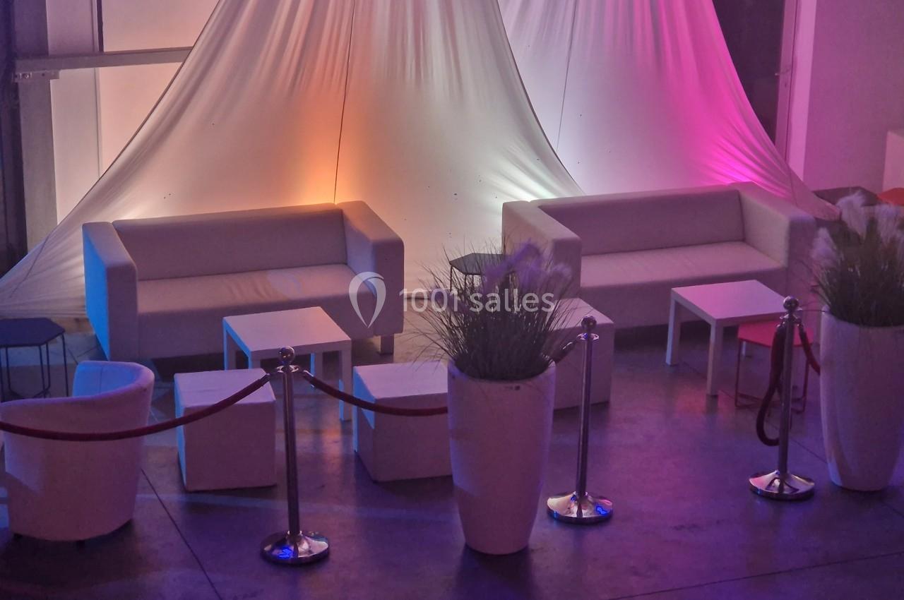 Espace lounge avec canapés blancs, tables basses et plantes décoratives, éclairé par des lumières colorées.