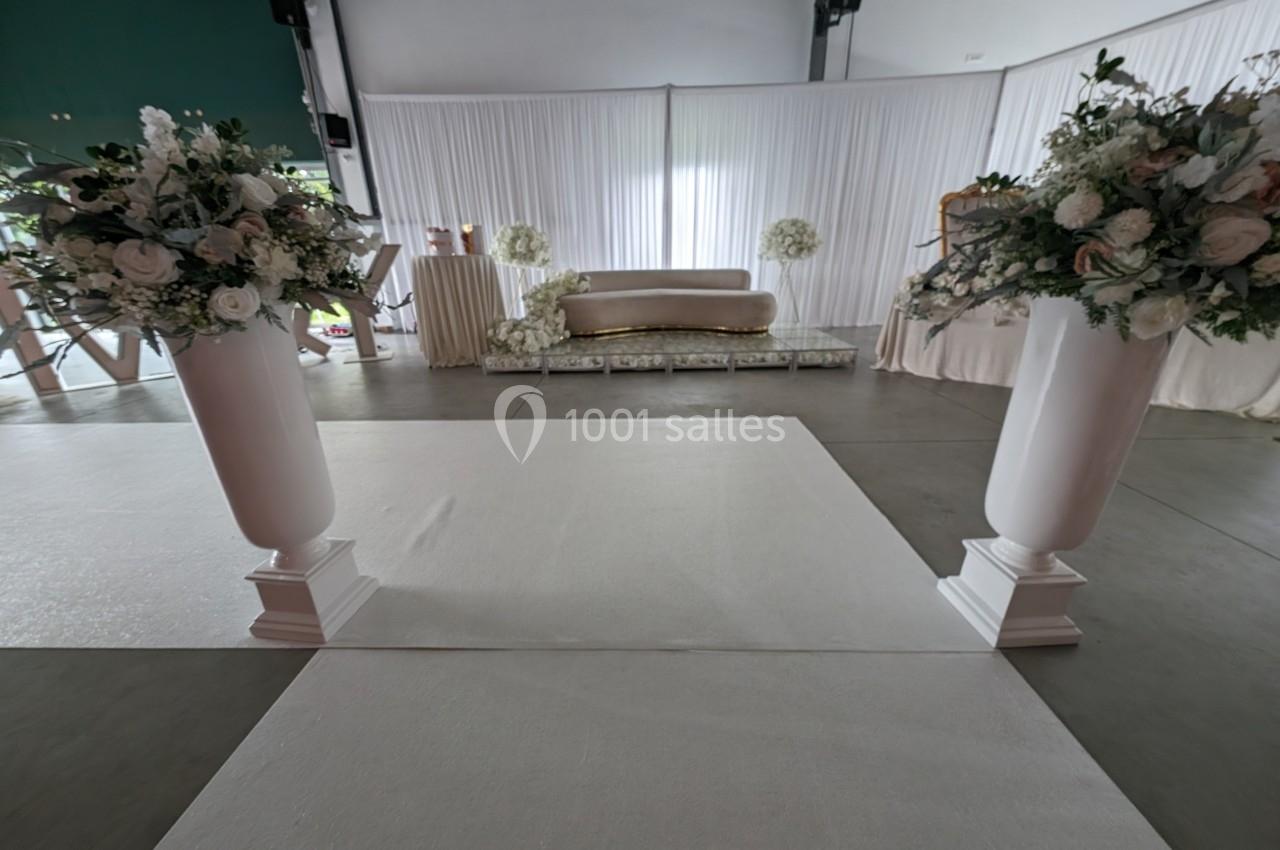 Salle décorée pour un événement avec des colonnes florales blanches, un canapé doré et des rideaux blancs en arrière-plan.