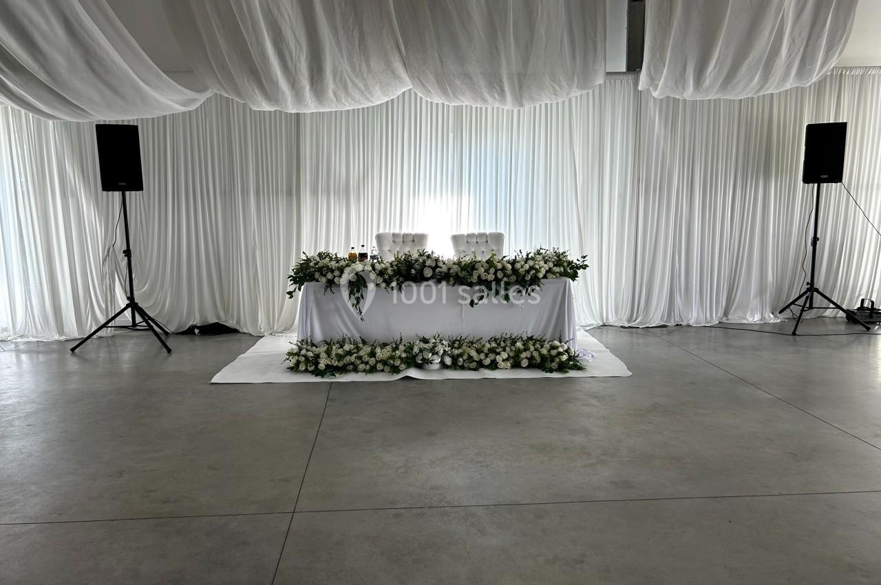 Table décorée de fleurs blanches et feuillages, placée sous des drapés blancs dans une salle au sol en béton.