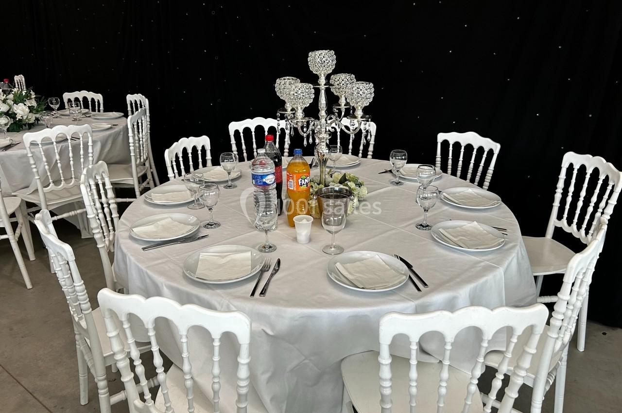 Table ronde dressée avec nappe blanche, chaises blanches, chandelier au centre, boissons et vaisselle disposées.