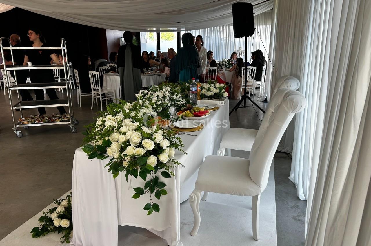 Table décorée de fleurs blanches dans une salle de réception, avec des invités assis en arrière-plan.