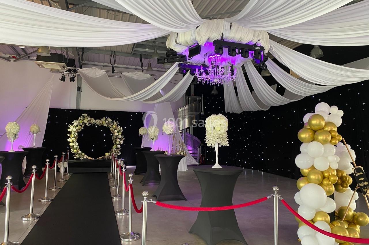 Salle de réception décorée avec des drapés blancs, un lustre, des ballons dorés et blancs, et une arche florale.
