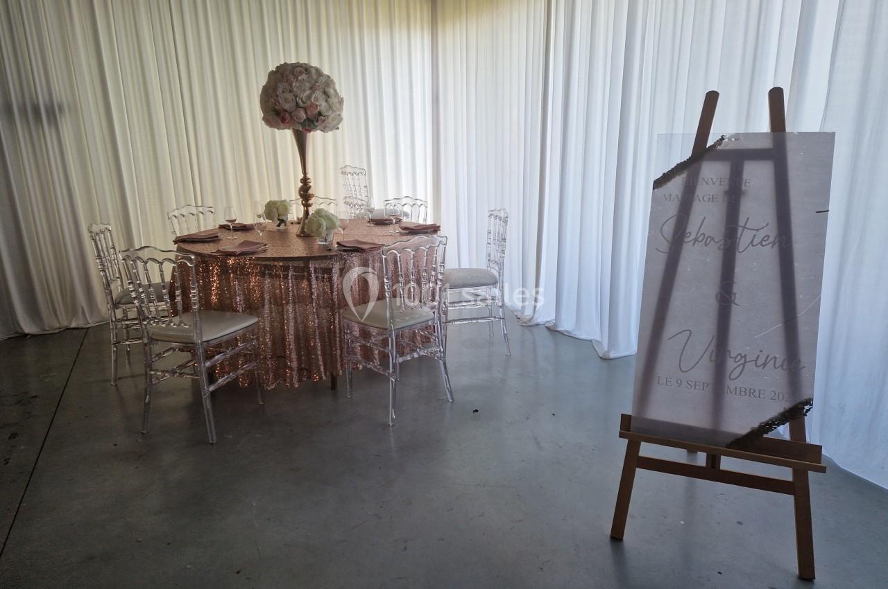 Table décorée pour un mariage avec nappes roses, chaises transparentes et centre de table floral, panneau d'accueil à droite.