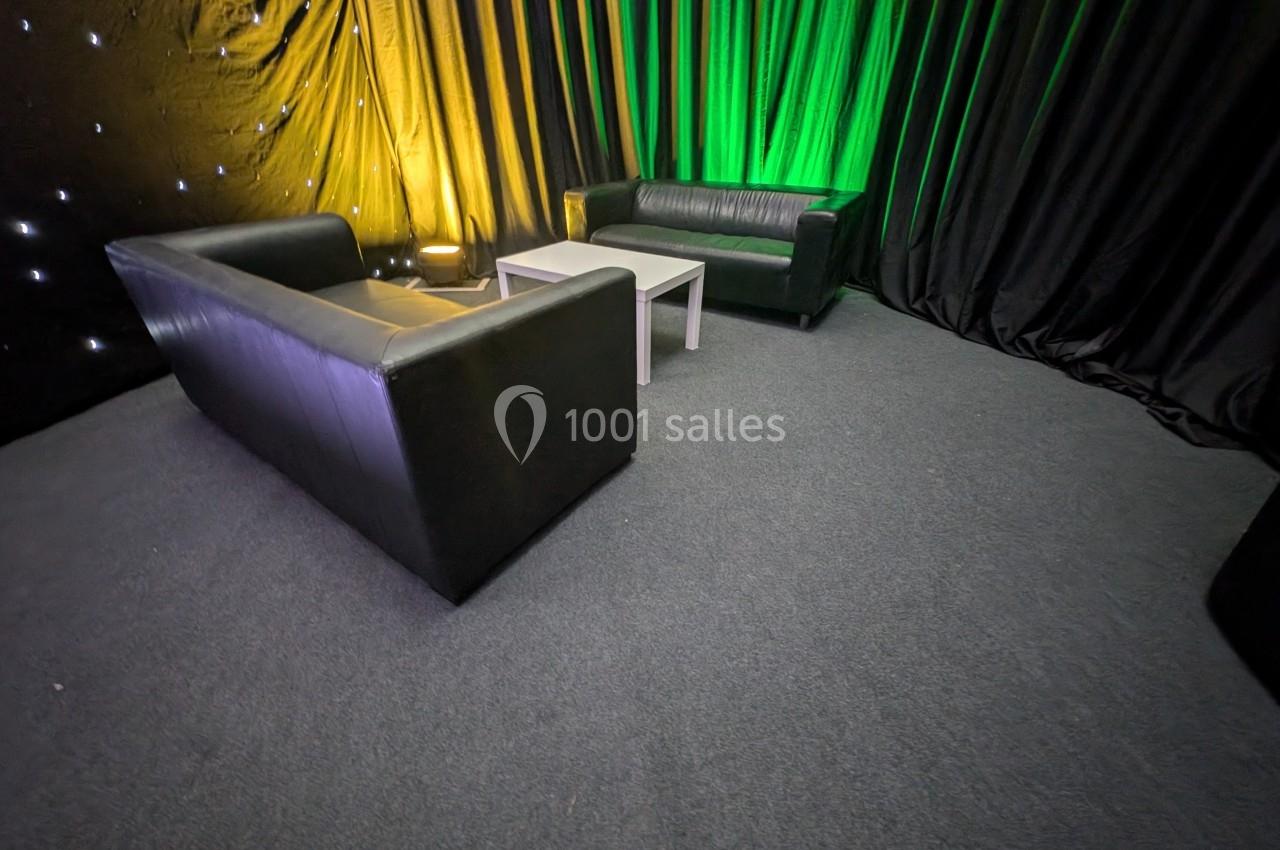 Deux canapés noirs entourent une table basse blanche dans une pièce décorée de rideaux noirs et éclairée en vert et jaune.