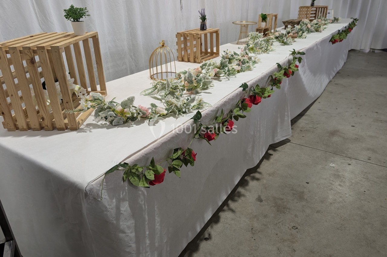 Table décorée avec des fleurs, feuillages, cages dorées et caisses en bois, sur une nappe blanche dans une salle.