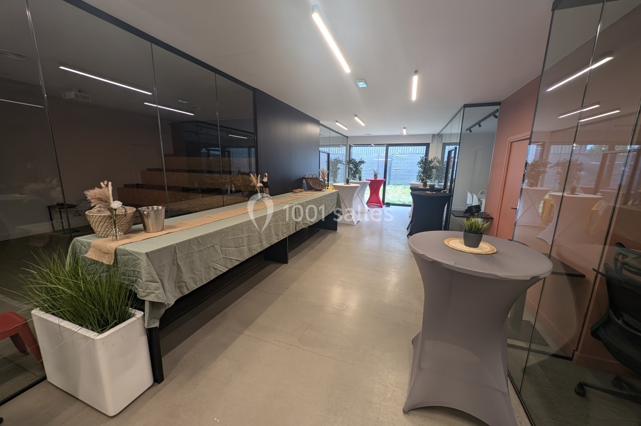 Salle moderne avec tables recouvertes de nappes, décorations simples et baies vitrées donnant sur un espace extérieur.