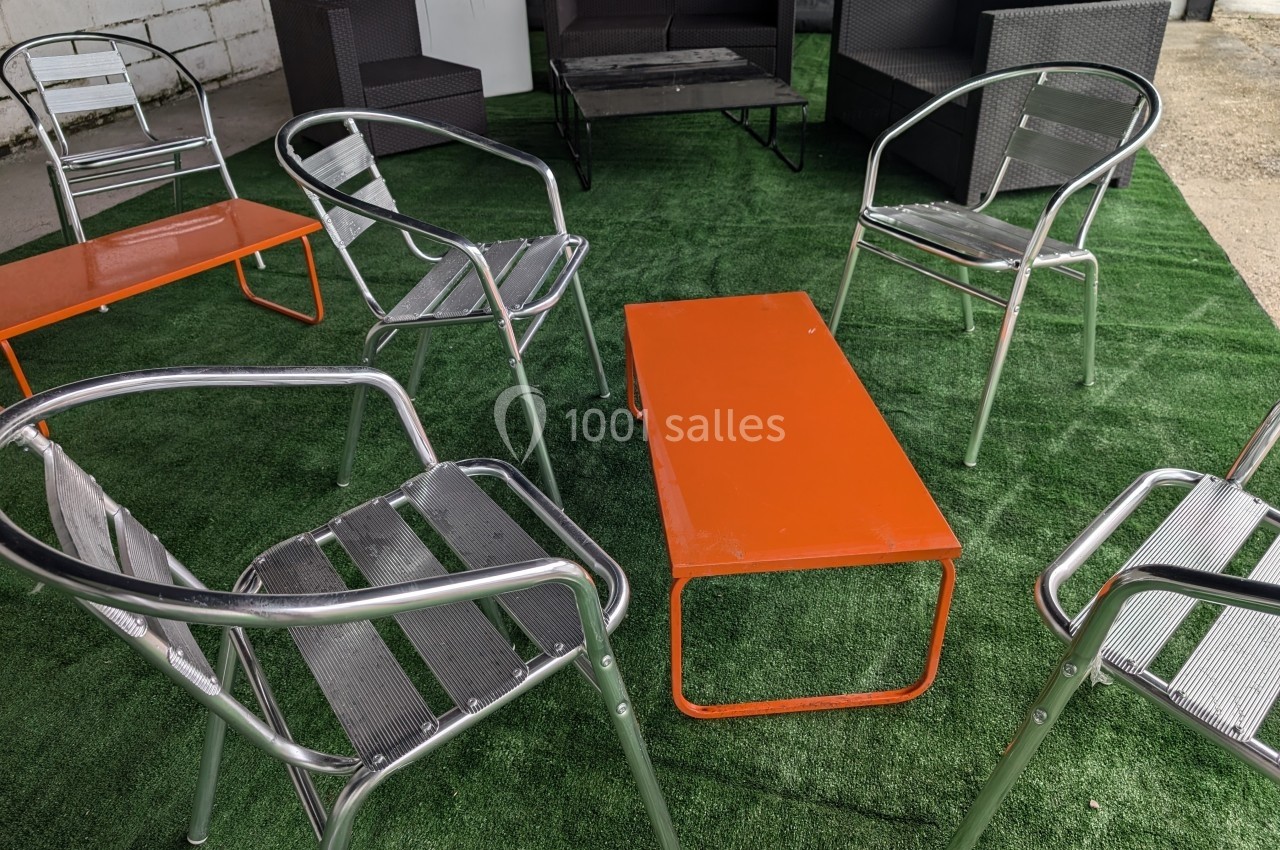 Chaises en métal et table orange disposées sur un tapis vert en extérieur, avec des canapés en arrière-plan.