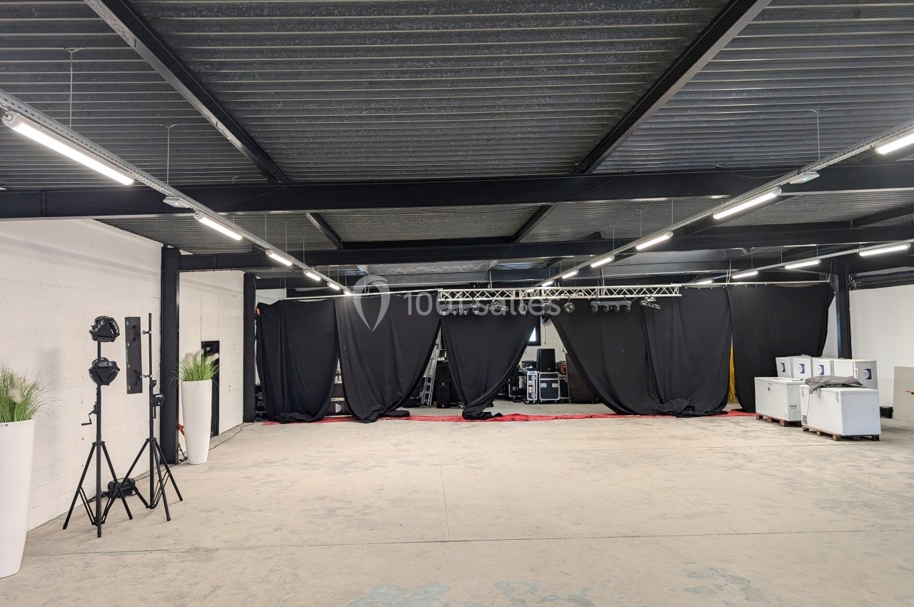 Salle vide avec sol en béton, plafond métallique, rideaux noirs couvrant le fond et équipements techniques visibles.