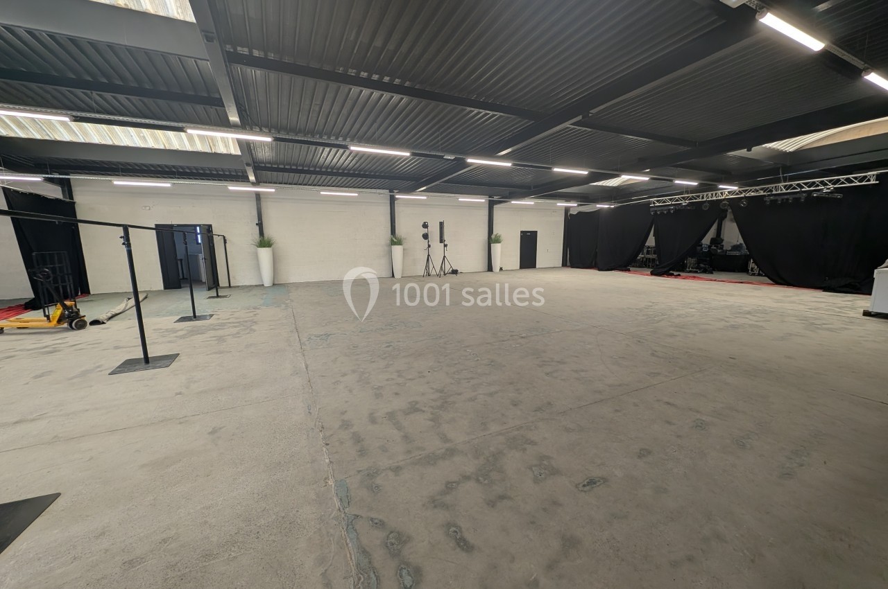 Salle spacieuse avec sol en béton, murs blancs, rideaux noirs et équipements techniques en arrière-plan.