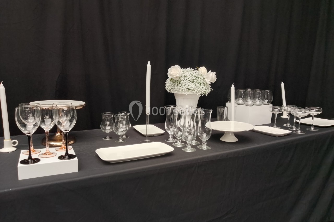 Table dressée avec des verres, des assiettes blanches, des bougies et un bouquet de fleurs blanches sur fond noir.