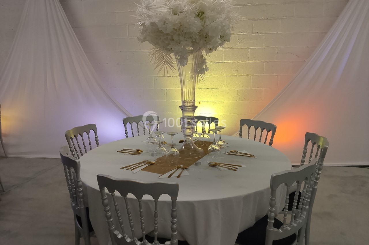 Table ronde décorée avec une nappe blanche, un centre de table floral et des chaises argentées, dans une salle éclairée.