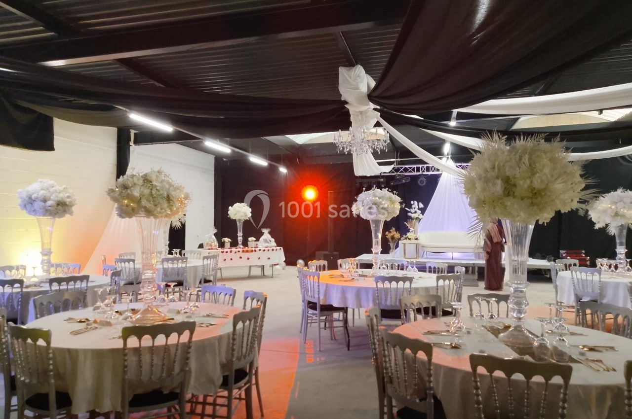 Salle de réception décorée avec des tables rondes, des chaises argentées, des centres de table fleuris et un éclairage…