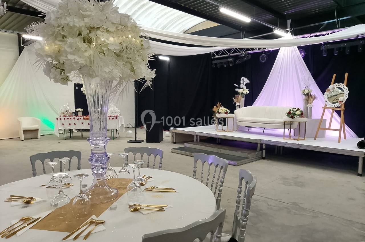 Salle de réception décorée avec une table dressée au premier plan et une scène ornée de fleurs blanches en arrière-plan.