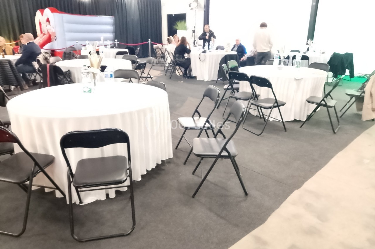 Salle avec des tables rondes couvertes de nappes blanches, entourées de chaises noires, et quelques personnes présentes.