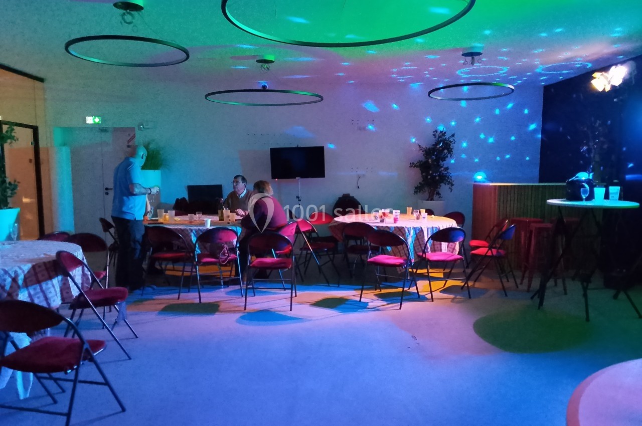 Salle éclairée par des lumières colorées, avec des tables, des chaises et quelques personnes discutant.