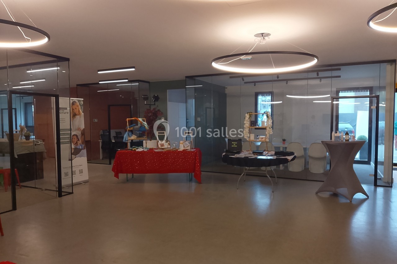 Salle avec des stands d'exposition, tables décorées et panneaux en verre, éclairée par des luminaires circulaires.