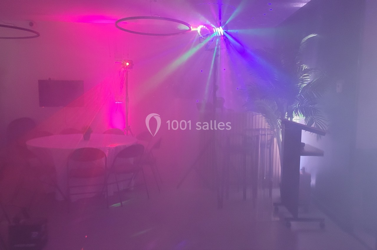 Salle faiblement éclairée avec des lumières colorées, des tables rondes et un pupitre, ambiance festive avec fumée.