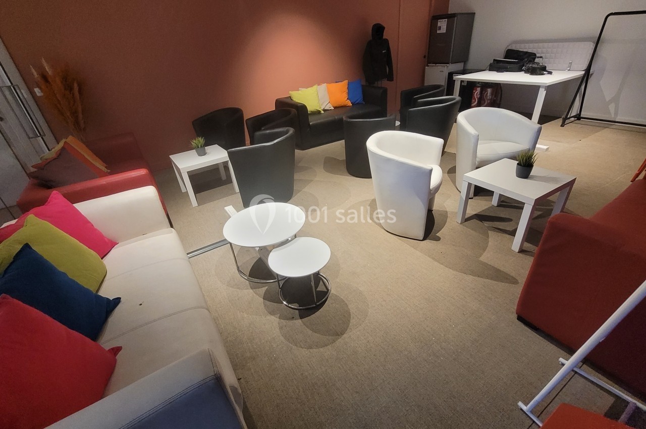 Salle aménagée avec des fauteuils, canapés colorés, tables basses blanches et noires, sur un sol en moquette beige.